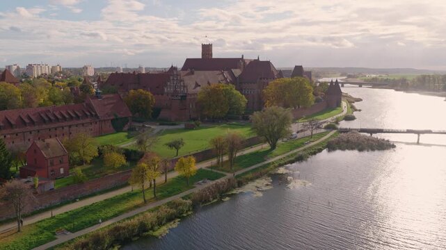 Aerial view of Malbork Castle in Malbork, Poland, with towers, walls, courtyards and riverside path. Camera tracks by bridges, water reflections and people in warm light.