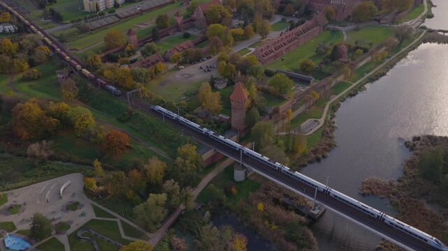 Aerial drone glide over Malbork Castle, towers and ramparts by the Nogat River, tiled roofs and parks, then a railway bridge with passing trains in late day light.