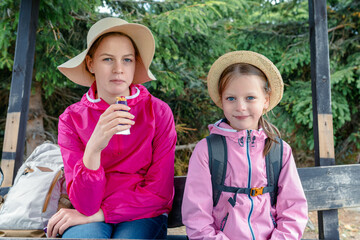 Two girls taking a snack break during a hike, sitting on a wooden bench in the forest wearing sun...