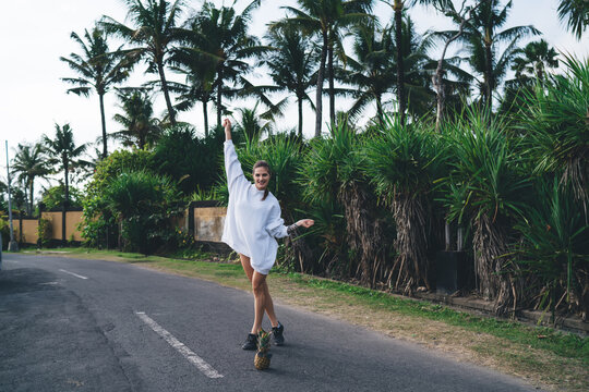 Smiling young woman joyfully walking down empty tropical road in oversized sweatshirt with pineapple, celebrating lightness, freedom and movement in remote digital lifestyle - Powered by Adobe