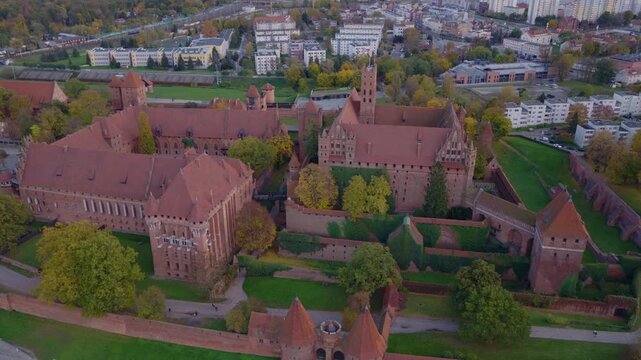 Aerial view of Malbork Castle in Poland reveals red brick halls, towers, walls, and gatehouse, with courtyards, ivy, paths, visitors, and late day light.