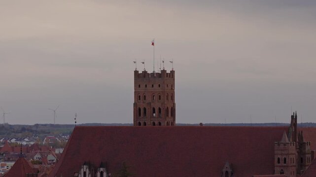 Aerial camera rises and sweeps over Malbork Castle in northern Poland, showing brick keep with flag, red tiled roofs, arches, battlements, town, turbines, and autumn trees.