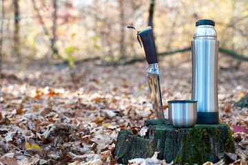 Obraz premium Thermos and hunting knife. The concept of tourism, survival and hunting in the autumn in forest. a thermos of tea, a cup and a knife, on an old tree stump. dry leaves in the park. at nature. close-up