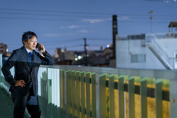街の夜景を背に緊急対応するサラリーマン、残業帰りに取引先へ連絡する男性社員 © hiro
