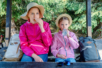 Girls resting on a forest bench during a hike, wearing sun hats and pink jackets, one eating a...