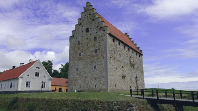 The camera panning across the well preserved medieval fortified manor of Glimmingehus, revealing the stone keep, moat, and bridge under a blue cloudy sky in the Skane region of Sweden
