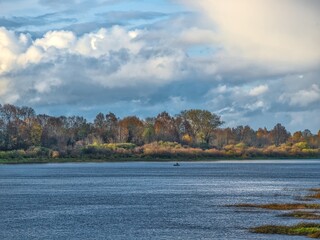 A bright, vast landscape of a wide river, with the opposite bank covered in a forest showing the vibrant and subtle colors of late autumn under a powerful, partly sunny, cloudy sky