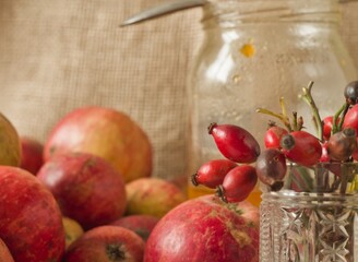 Glass jar and vase of rose hips next to apples in a muted, rustic still life