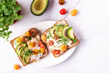 Tasty homemade toasts for breakfast with fried eggs, cream cheese, avocado, cherry tomatoes and fresh oregano leaves on white marble background top view.
