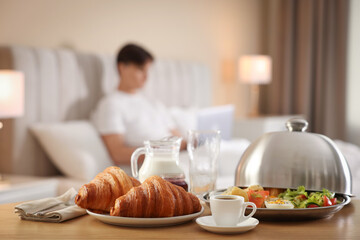 Delicious breakfast served on wooden table and man working with laptop in bed at hotel room, selective focus