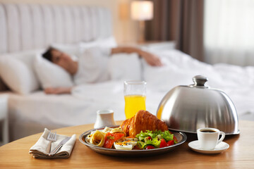 Delicious breakfast served on wooden table and man sleeping in bed at hotel room, selective focus