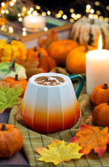 An orange white cup of coffee latte with Halloween ghost draw with cacao powder as latte art, autumn leaves and assorted Jack-Be-Little squash, fall ambiance with bokeh light and candles in background