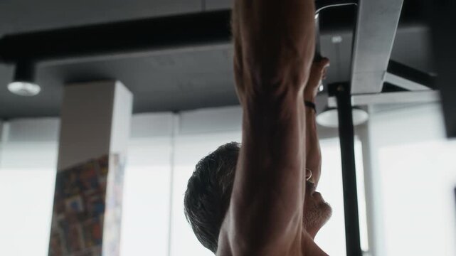 Shirtless senior Caucasian man with strong body doing pull-up exercise in modern gym, low angle view