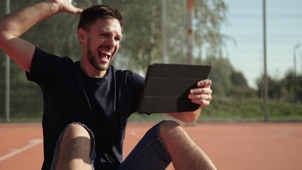 Caucasian man sits on outdoor sports field, emotionally celebrating favorite team victory. Fan watches sports match on tablet and celebrates winning bet. - Powered by Adobe