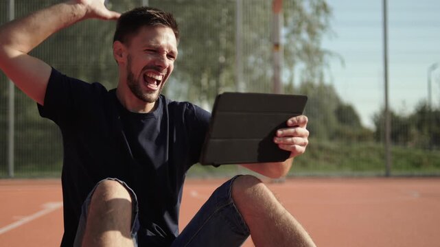 Caucasian man sits on outdoor sports field, emotionally celebrating favorite team victory. Fan watches sports match on tablet and celebrates winning bet.