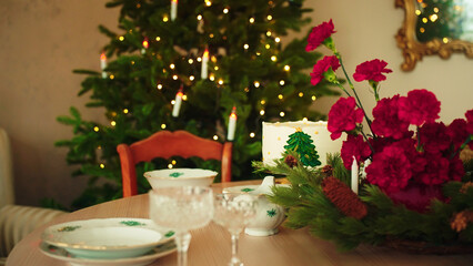 Cozy holiday table with white cake, red carnations, pine branches, and vintage porcelain. Blurred Christmas tree in background. Perfect for marketing with copy space. Christmas background