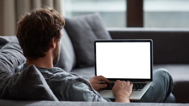 Young male tech user relaxing on sofa holding laptop computer mock up blank white screen. Man using modern notebook surfing internet, read news, distance online study work concept. Over shoulder view