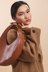 asian woman wearing a warm brown coat and carrying a curved leather bag poses against a soft...