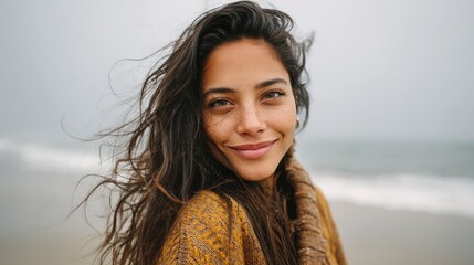 Portrait of young woman at sea looking at camera. Smiling latin hispanic girl standing at the beach with copy space and looking at camera. Happy mixed race girl in casual outfit with wind in her hair