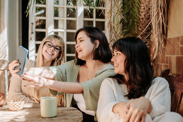 Three women taking selfies and laughing together outdoors, enjoying coffee and friendship in a warm, natural setting with soft light and cozy tones.