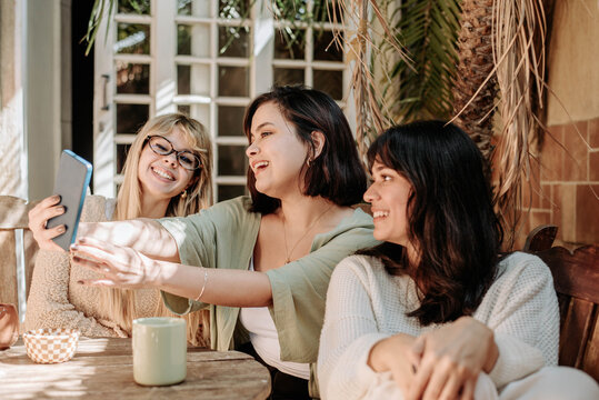 Three women taking selfies and laughing together outdoors, enjoying coffee and friendship in a warm, natural setting with soft light and cozy tones.