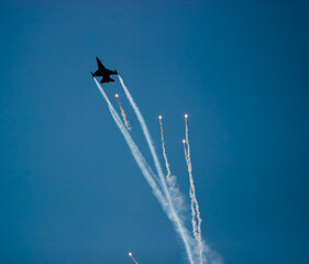 A fighter plane is seen throwing heat flares (flare) in the blue sky background. Bright light traces and smoke clouds reveal the dynamism and visual appeal of the maneuver.