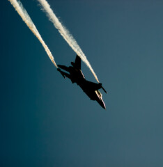 A fighter plane, which makes a turning maneuver against sunlight, leaves traces of intense smoke behind. The sharp silhouette in the sky emphasizes the agility and speed of the plane.