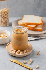 Creamy peanut butter in a jar on a wooden plate on a blue background with a scattering of roasted nuts