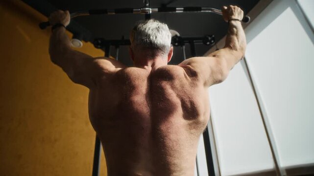 Back view of shirtless mature man with muscular body and gray hair doing lat pulldowns in modern gym