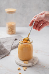 A hand holds a spoon with creamy peanut butter in a jar on a light marble background