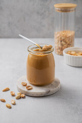 Creamy peanut butter in a jar on a light background with roasted nuts and spoon.
