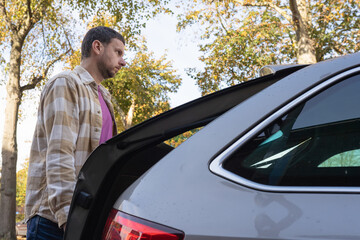 Man with short hair wearing a plaid shirt and pink t-shirt is standing beside a car trunk in a park, surrounded by trees and autumn foliage, enjoying a moment outdoors
