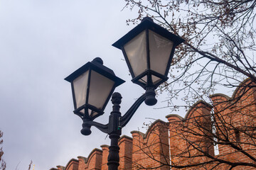 Vintage black forged street lamp against an old red brick fortress wall with battlements. Urban background for the concept of history, security, and city street illumination.
