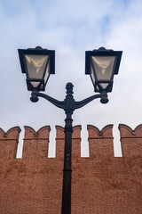 Vintage black forged street lamp against an old red brick fortress wall with battlements. Urban background for the concept of history, security, and city street illumination.