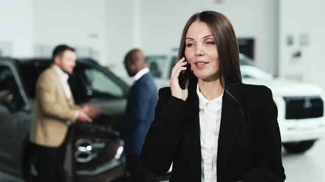A young woman talks on the phone and smiles in a modern car dealership, while in the background a dealer and a customer finalize a deal. Concept buying a new car.