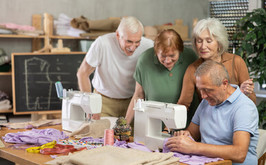 Elderly students watch senior man teacher work with sewing machine during lesson at sewing school....