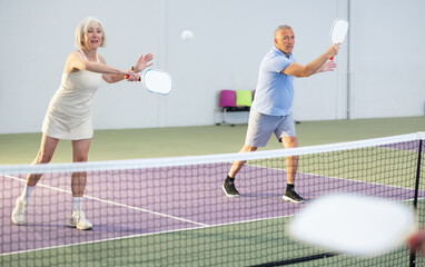 Obraz premium Sporty couple of senior woman and man playing doubles pickleball on indoor court, ready to hit ball. Sport and active lifestyle concept..