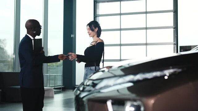 An African American man in a blazer shakes hands with a Caucasian woman at a car dealership, completing the purchase of a new car. Concept buying a new car.