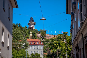 Obraz premium Graz, Austria – July 22, 2025: The iconic Uhrturm (Clock Tower) on Schlossberg Hill stands tall above vibrant gardens, under a moody summer sky in the historic heart of Graz. 