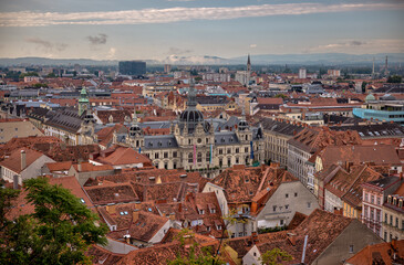 Obraz premium Graz, Austria – July 22, 2025: A scenic panoramic view of the historic city center with red rooftops, church spires, and lush summer foliage under a dramatic morning sky. 