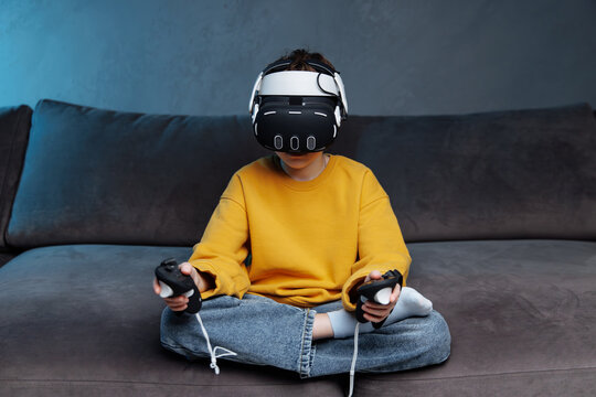 A young Caucasian boy of 10 years old wearing a virtual reality helmet, sitting on a couch with game controllers in his hands, immersed in the gameplay in augmented reality
