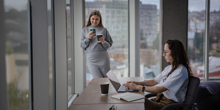 Two women work in an office. The girl is working on a laptop, the second woman - pregnant - is working from the phone, standing by the window