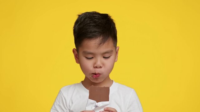 A young Asian boy savoring a chocolate bar with a thoughtful expression. Bright yellow background enhances the cheerful atmosphere, capturing a moment of childhood joy and indulgence.