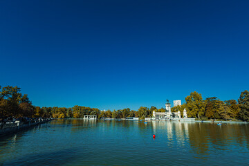 Obraz premium A calm lake reflects the grand monument at El Retiro Park in Madrid. People relax on steps and row small boats under a clear blue sky, surrounded by trees in warm seasonal colors.