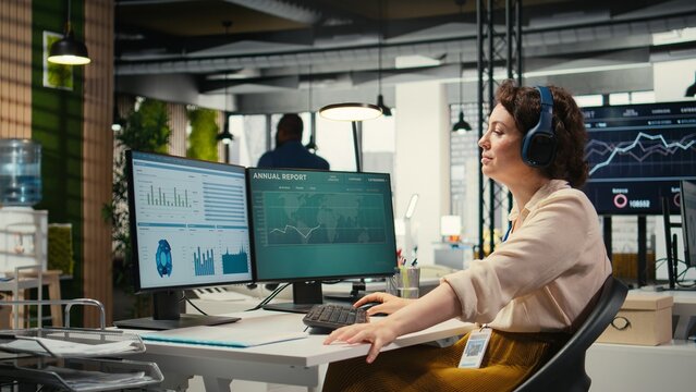 Young businesswoman focused on a podcast while she resolves office tasks, monitoring insights or KPI metrics on pc. Strategy reports for expansion plans with digital tools.