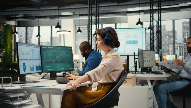 Office administrator uses focus music on headset to solve tasks, monitoring performance metrics at the workstation. Administrative tasks to ensure enterprise productivity and success.