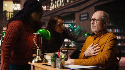 Elderly college student does inhale exhale exercise due to physical discomfort, having a respiratory problem in distress at the library desk. Aged man unwell in distress.