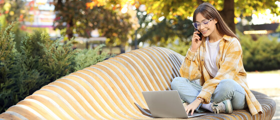 Young woman with laptop talking by mobile phone on bench outdoors