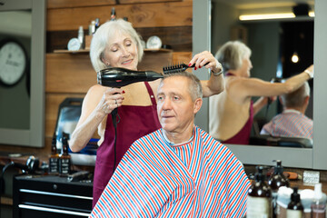 Elderly woman hairdresser completes creation of haircut and dries hair with hairdryer to elderly...