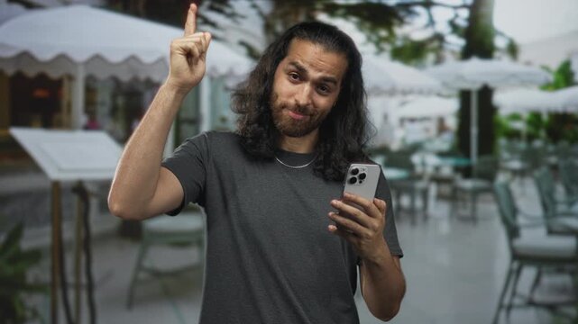 Man points finger up holding smartphone and smiling with raised hand on street terrace cafe outdoors; confidence quick tip.